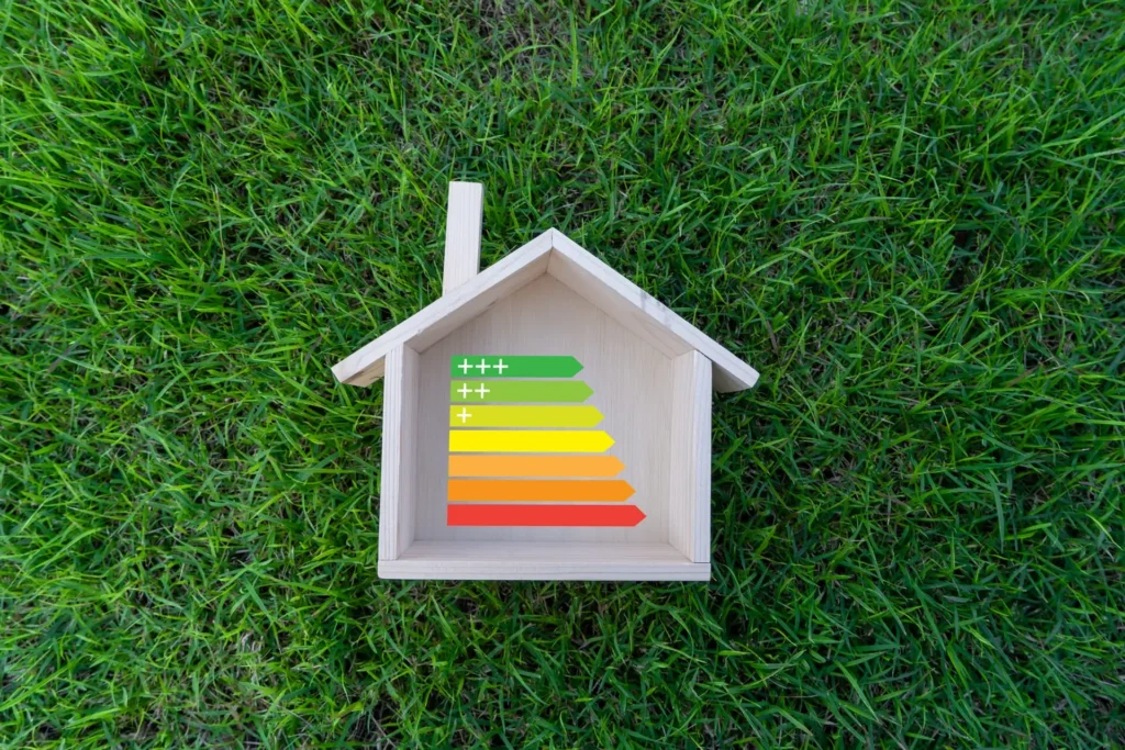 Uma casinha de madeira em miniatura está sobre a grama verde. Dentro dela, um gráfico colorido em barras mostra os níveis de eficiência energética - do verde (mais eficiente) ao vermelho (menos eficiente). A imagem destaca como é importante entender o que é kVA para o consumo de energia em residências, tema fundamental para a Tecnogera.