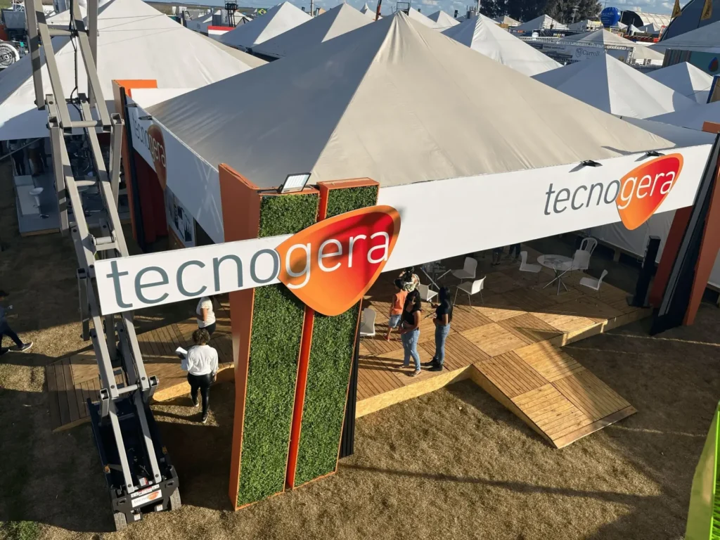 No estande da Tecnogera na Bahia Farm Show, grandes tendas brancas e uma placa verde e laranja com o nome da empresa chamam a atenção. Os visitantes circularam pelo deque de madeira sobre a grama seca, conhecendo as oportunidades do agronegócio. Mesas e cadeiras ficam dispostas sob as tendas, enquanto o sol forte ilumina o espaço e projeta sombras nítidas ao redor.