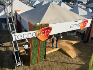 No estande da Tecnogera na Bahia Farm Show, grandes tendas brancas e uma placa verde e laranja com o nome da empresa chamam a atenção. Os visitantes circularam pelo deque de madeira sobre a grama seca, conhecendo as oportunidades do agronegócio. Mesas e cadeiras ficam dispostas sob as tendas, enquanto o sol forte ilumina o espaço e projeta sombras nítidas ao redor.