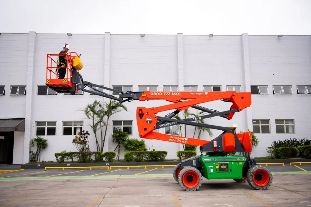 Um colaborador, equipado com itens de segurança, opera uma Plataforma Elevatória da Tecnogera ao lado de um prédio industrial branco com janelas e plantas. A plataforma está assentada sobre uma área de estacionamento demarcada por linhas amarelas, sob um céu nublado.