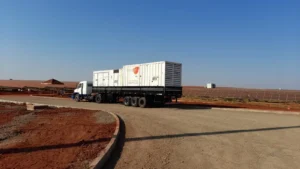 Um caminhão branco com carreta prancha transporta dois grandes contêineres brancos e está estacionado em uma estrada de terra e cascalho. Ao fundo, há fileiras de painéis solares instaladas sobre o solo avermelhado, reforçando o compromisso da Tecnogera com soluções de energia para a gestão do agronegócio sob um céu azul claro.
