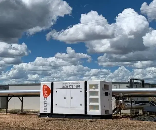 Um grande gerador de energia branco da Tecnogera, posicionado sobre cascalho próximo a tubulações industriais, se destaca sob um céu azul. O equipamento exibe o telefone de contato, sendo perfeito para quem busca comprar ou alugar geradores nas proximidades do edifício cercado.