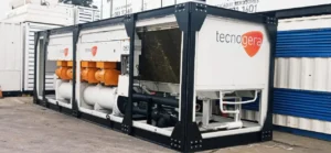 Um grande chiller industrial da Tecnogera, com o nome da marca em laranja, está instalado ao ar livre. O equipamento conta com tubulações e painéis de controle e foi desenvolvido para garantir um controle eficiente de temperatura em operações industriais. Ao fundo, é possível ver contêineres de transporte.