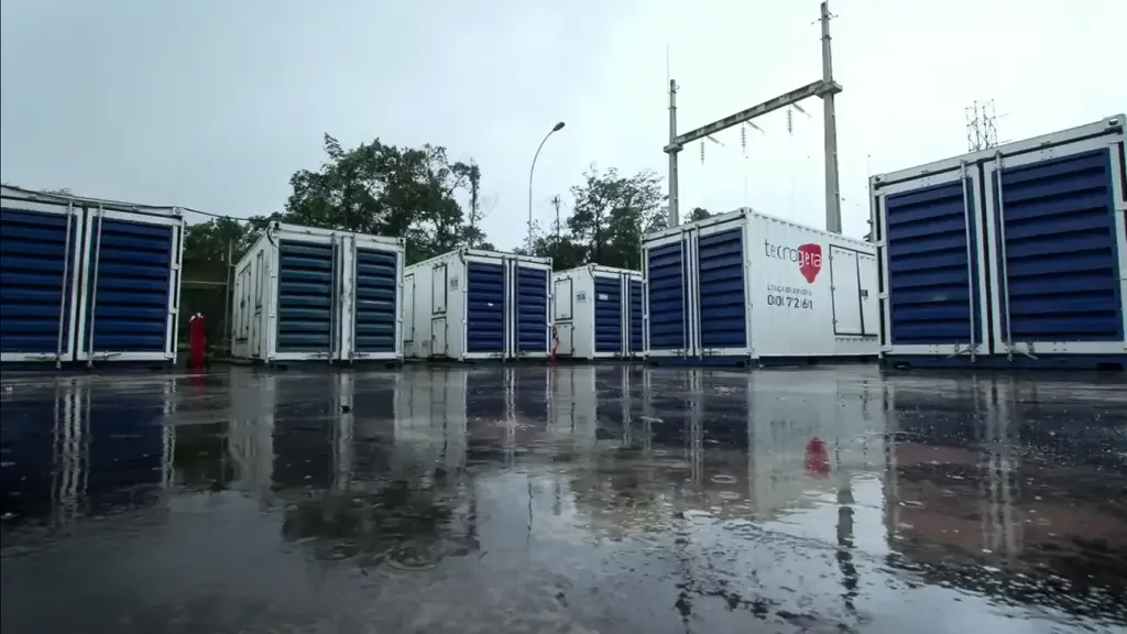 Vários grandes contêineres brancos e azuis de baterias industriais da Tecnogera, usados no setor de mineração, estão posicionados ao ar livre sobre um piso molhado e refletivo. O cenário mostra poças d'água causadas pela chuva recente, com árvores e postes ao fundo sob um céu nublado.