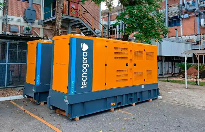 Dois grandes geradores industriais, nas cores laranja e azul e com o logotipo da Tecnogera, estão posicionados ao ar livre próximos a uma instalação de empresa estatal. Ao fundo, é possível ver árvores, áreas pavimentadas e estruturas do setor de Óleo & Gás.