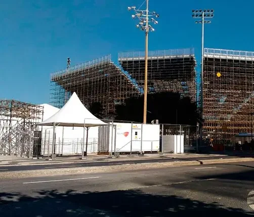Uma grande arquibancada metálica, parte da construção para as Olimpíadas Rio 2016, destaca-se sob um céu azul e sem nuvens. Na frente dela, vemos tendas brancas, um prédio móvel, cercas e uma estrada com faixas amarelas. Altos postes de luz completam o cenário ao redor da estrutura. Esta é mais uma operação que conta com a expertise da Tecnogera.