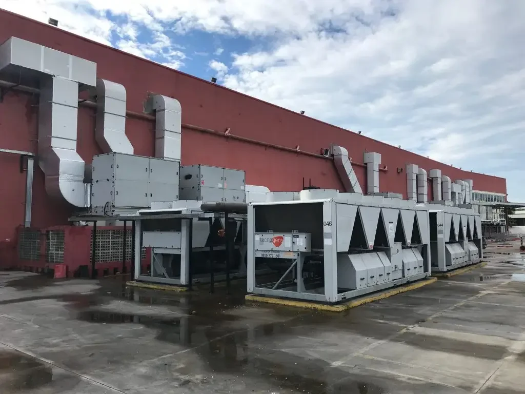 Unidades industriais de HVAC e dutos de ventilação de grande porte estão instalados do lado de fora de um prédio industrial vermelho, parecido com uma megaloja. O chão de concreto molhado reflete os equipamentos, enquanto grandes ventilações e tubulações metálicas se estendem até o edifício sob um céu parcialmente nublado. Tudo isso faz parte das soluções modernas oferecidas pela Tecnogera.