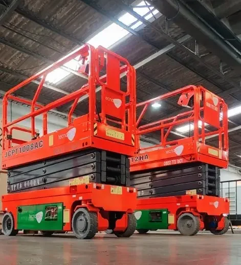 Duas plataformas elevatórias tipo tesoura nas cores laranja e preto da Tecnorent estão estacionadas em um ambiente interno, sobre um piso de concreto liso, sob um amplo teto industrial com claraboias. Ambas as plataformas, perfeitas para locação pela Tecnogera, estão levemente elevadas e contam com graus de segurança ao redor.