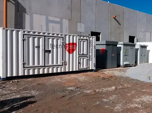 Um contêiner branco com o logotipo vermelho da Tecnogera, utilizado para soluções de colocation da Odata, está ao ar livre ao lado de um prédio industrial cinza. Próximos a ele, há dois equipamentos elétricos cinzas sobre um terreno irregular e sem pavimentação, sob um céu azul e aberto.