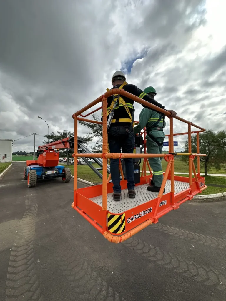 Dois trabalhadores, equipados com cintos de segurança, estão sobre uma plataforma elevatória laranja identificada com a inscrição "CAPACIDADE 230kg", em uma área externa asfaltada. A plataforma está suspensa e, ao fundo, é possível ver um grande equipamento do tipo boom lift sob um céu nublado. Essa é uma das soluções oferecidas pela Tecnogera para trabalho seguro em altura.