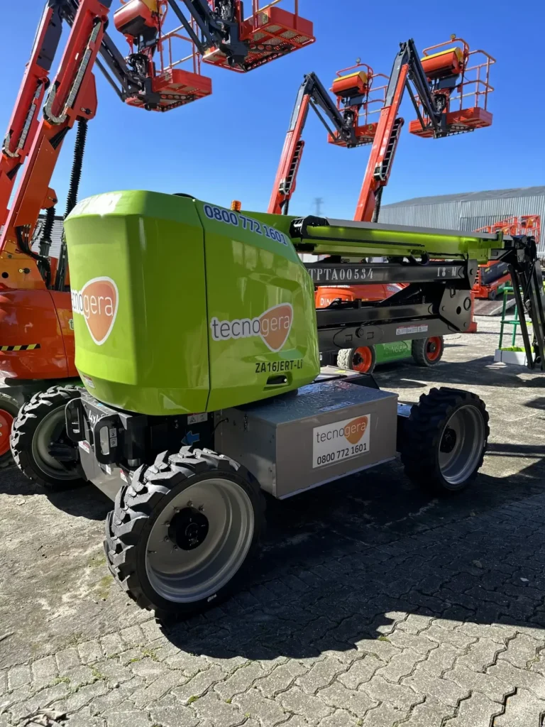 Uma plataforma elevatória telescópica verde, com a marca Tecnogera, está estacionada em um pátio pavimentado e com o braço abaixado, pronta para contribuir com a produtividade. Ao fundo, sob o sol forte, aparecem outros equipamentos do tipo boom lift nas cores laranja e preto sobre blocos de concreto intertravados.