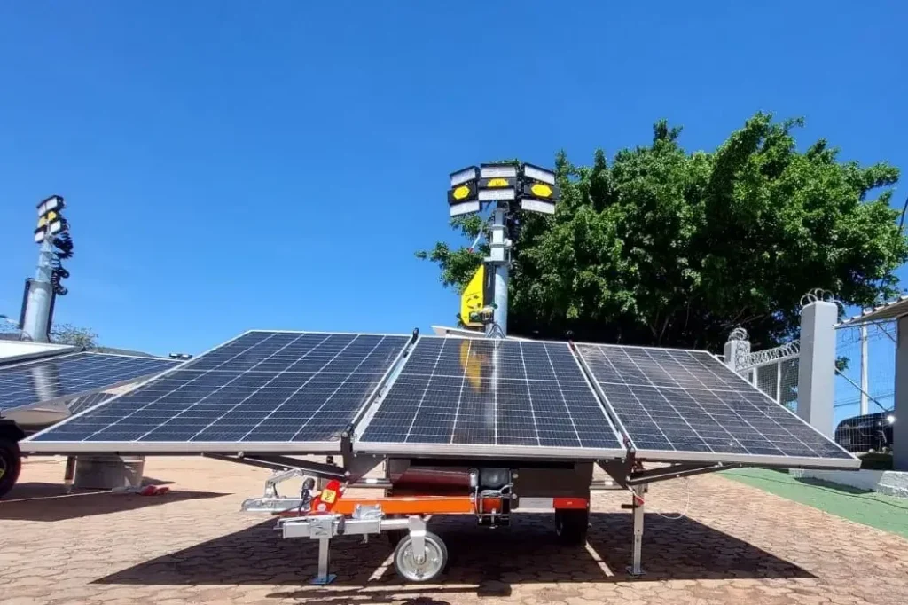 Um conjunto móvel de painéis solares sobre rodas está ao ar livre sob um céu azul, inclinado para captar o máximo de luz do sol. Perto dali, uma torre de iluminação solar da Tecnogera funciona ao lado de uma grande árvore frondosa e uma área cercada.