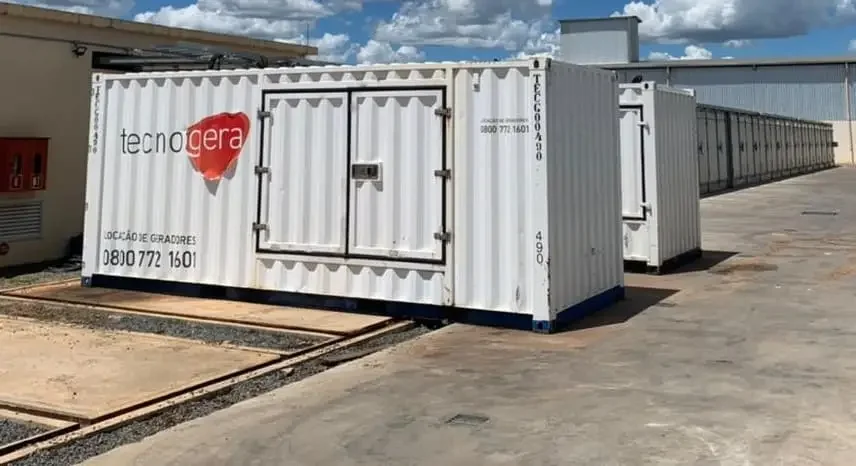 Uma fileira de grandes contêineres brancos, com o logotipo vermelho e cinza da Tecnogera e informações de contato, está posicionada sobre uma área de concreto. Os equipamentos estão disponíveis para aluguel de gerador de energia em frente a um prédio industrial, sob um céu parcialmente nublado.