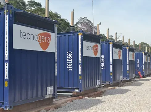 Diversos contêineres azuis com a marca "Tecnogera" estão alinhados sobre um caminho de cascalho, compondo parte de uma usina termelétrica. Cada contêiner possui um número de identificação e hastes verticais em seu topo. Ao fundo, é possível ver árvores e o céu nublado.