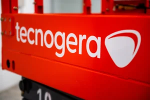 Close-up de uma máquina industrial vermelha, destacando o nome Tecnogera em branco e seu logotipo estilizado em formato de escudo. A imagem ressalta o compromisso da marca com energia em movimento, enquanto o fundo desfocado faz com que a atenção fique na cor vibrante e no visual da Tecnogera.