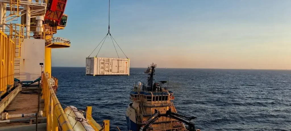 Um contêiner branco da Tecnogera está sendo içado por um guindaste de uma plataforma de petróleo offshore em direção a um navio de suprimentos, enquanto o mar tranquilo e o céu claro ao entardecer representam um novo momento no setor de energia.