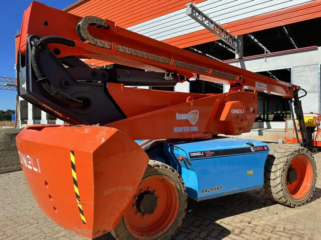 Imagem em close de uma plataforma elevatória articulada nas cores laranja e azul, identificada com a marca "Dingli", estacionada sobre um piso pavimentado em um canteiro de obras. O braço da máquina está levantado e, ao fundo, é possível ver um prédio com painéis vermelhos e brancos. Esta é mais uma solução eficiente da Tecnogera para trabalhos em altura.
