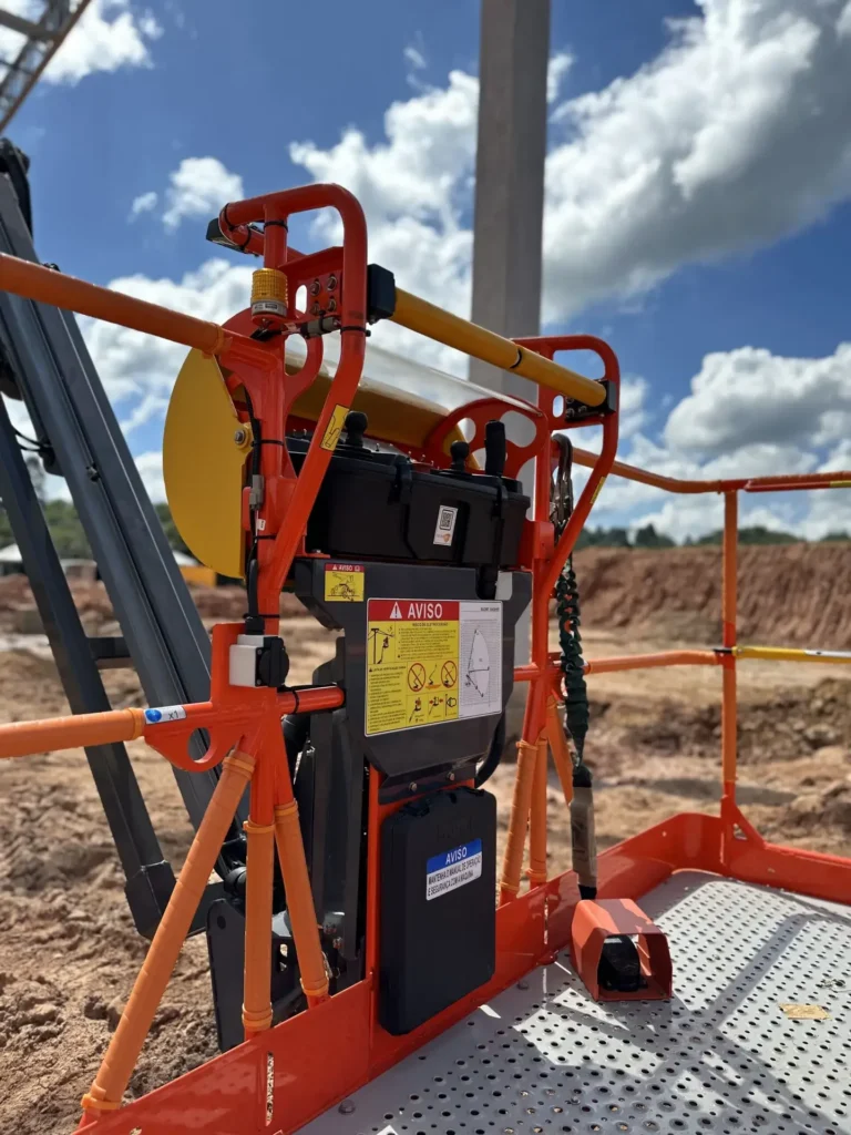 Imagem mostra um close-up de corrimão laranja e dos comandos de uma plataforma elevatória, com etiquetas de segurança em destaque. Ao fundo, vemos terra, materiais de construção e o céu azul, reforçando a importância dos cuidados ao alugar ou operar esse tipo de equipamento em obras movimentadas. A Tecnogera orienta sobre o uso seguro das soluções oferecidas.