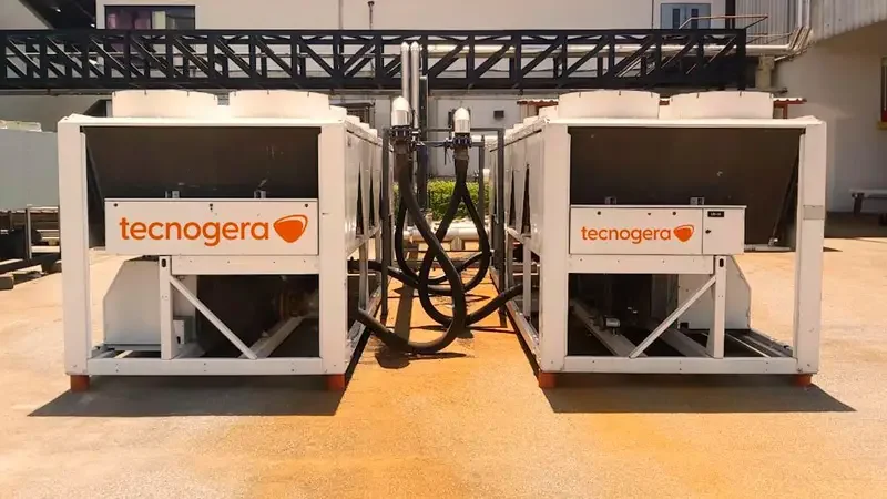 Dois grandes chillers industriais da Tecnogera estão instalados em uma área externa pavimentada, provavelmente atendendo a uma indústria de celulose. Mangueiras pretas conectam os equipamentos, e ao fundo é possível ver um prédio com uma passarela metálica.