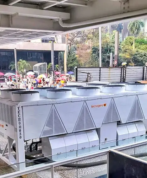 Unidades de ar-condicionado industrial de grande porte da Tecnofrio estão instaladas em uma área pavimentada ao ar livre, próxima a um prédio moderno. Plataformas para trabalho em altura da Tecnogera também aparecem no local. O cenário se completa com árvores, pessoas com guarda-chuvas coloridos e altas estruturas de vidro ao fundo.
