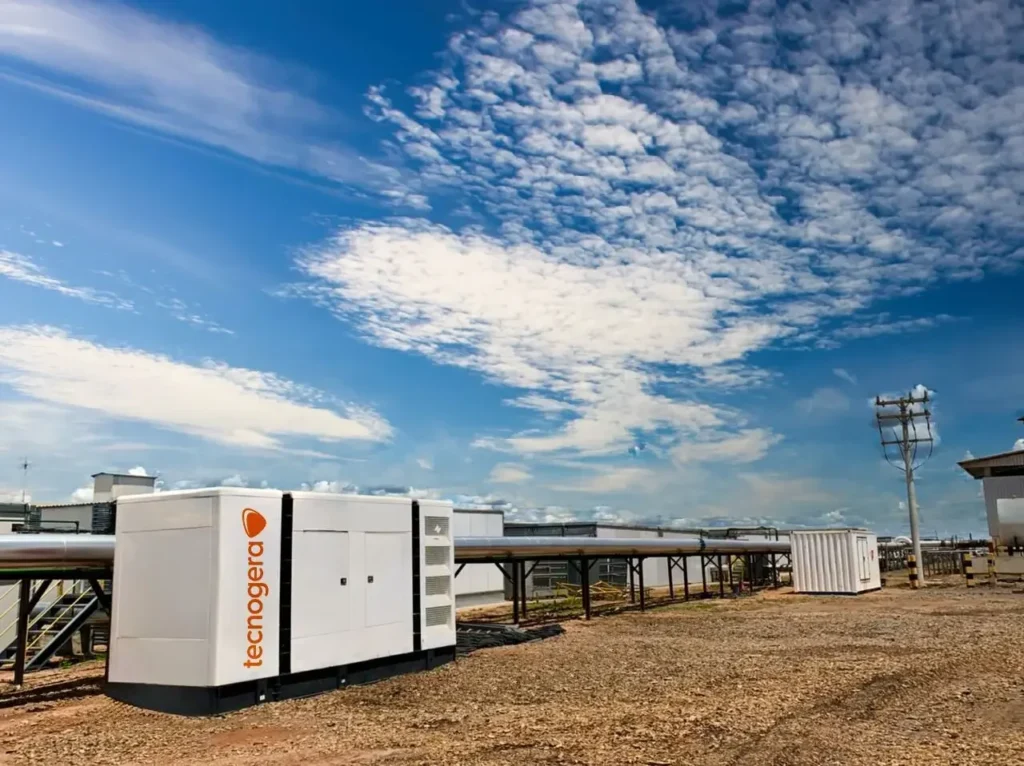 Um grande contêiner industrial branco, com o logotipo laranja da Tecnogera, está posicionado sobre um terreno de cascalho sob um céu azul brilhante. Ao fundo, é possível ver tubulações e estruturas metálicas se estendendo, além de linhas de energia à direita. O contêiner é utilizado para a locação de equipamentos de energia.