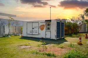 Um contêiner branco com a marca Tecnogera está posicionado sobre a grama, sob um céu de pôr do sol, ilustrando os desafios da organização de eventos: como garantir estrutura adequada e fornecimento contínuo de energia em um ambiente calmo e industrial. Pequenas árvores e extintores de incêndio completam o cenário ao redor.
