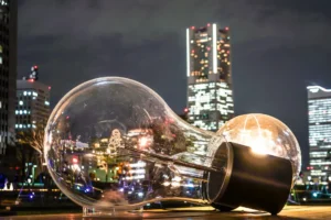 Uma grande escultura de lâmpada clara repousa de lado ao ar livre, em uma cidade iluminada à noite, refletindo os prédios brilhantes ao redor. Essa obra artística simboliza a gestão de energia e destaca como é fundamental ter um plano de contingência energética na movimentada paisagem urbana. É esse compromisso com soluções inteligentes que faz parte do dia a dia da Tecnogera.