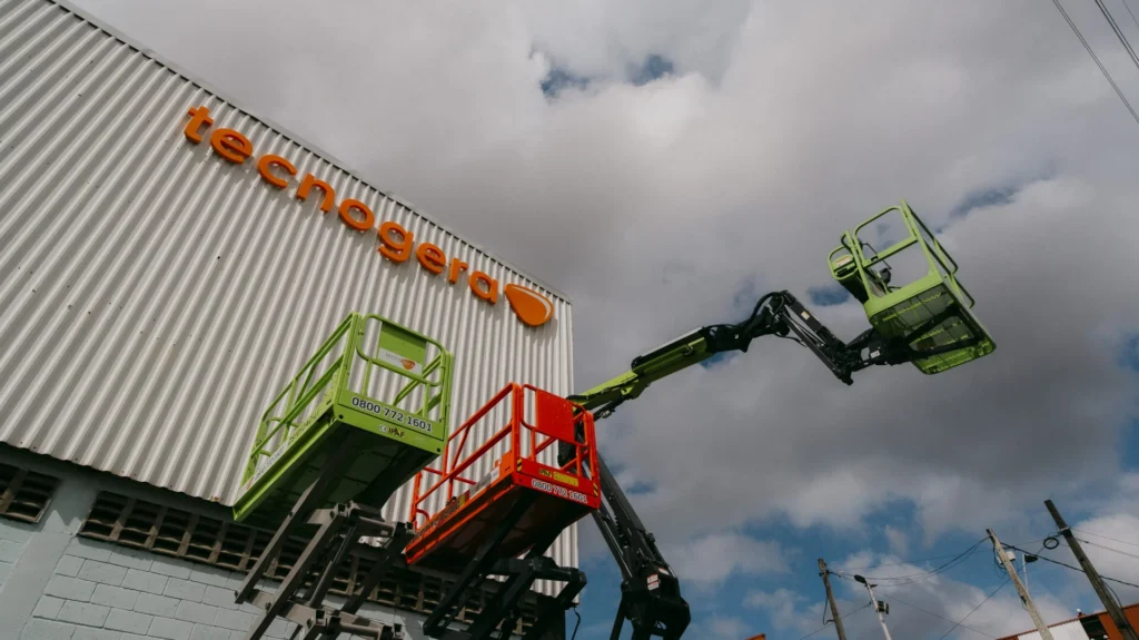 Duas plataformas elevatórias verdes alcançam um galpão de metal ondulado, que exibe o logotipo laranja da Tecnogera na fachada. O céu está nublado, fios elétricos aparecem ao fundo e uma das plataformas sustenta um cesto vermelho. Tecnogera: muito mais do que locação de geradores.