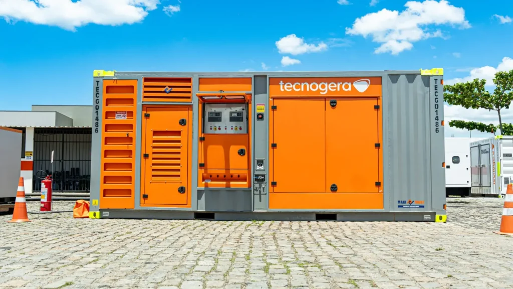 Um grande gerador cinza e laranja da Tecnogera está posicionado, garantindo segurança energética