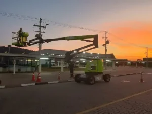 Um colaborador da Tecnogera, utilizando cinto de segurança conforme as normas NR35, manobra uma plataforma articulada verde ao lado de um prédio durante o pôr do sol. Cones laranjas sinalizam a área para garantir a segurança, enquanto postes, fios elétricos e um céu em tons vibrantes de laranja e azul compõem o cenário ao fundo