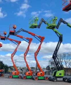 Plataformas elevatórias posicionadas em um pátio de cascalho sob um céu azul com nuvens, tendo ao fundo alguns prédios e árvores. Tudo isso faz parte da estrutura que a Tecnogera oferece para soluções eficientes em trabalhos em altura