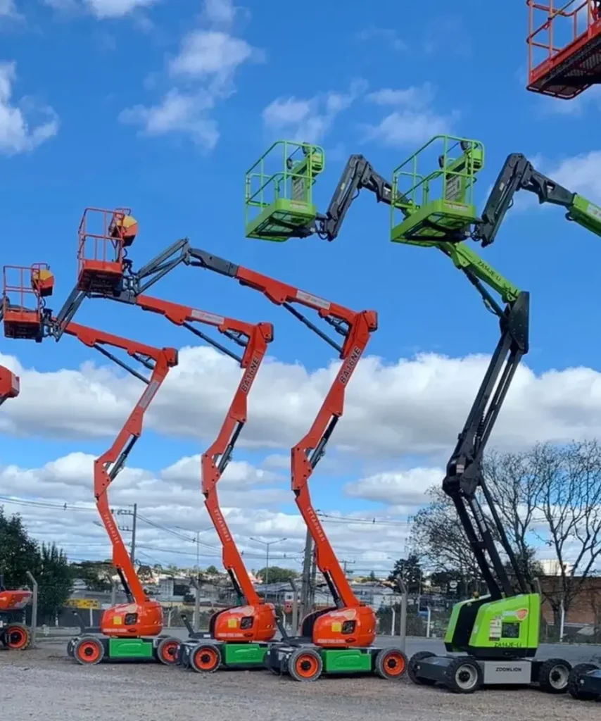 Plataformas elevatórias posicionadas em um pátio de cascalho sob um céu azul com nuvens, tendo ao fundo alguns prédios e árvores. Tudo isso faz parte da estrutura que a Tecnogera oferece para soluções eficientes em trabalhos em altura