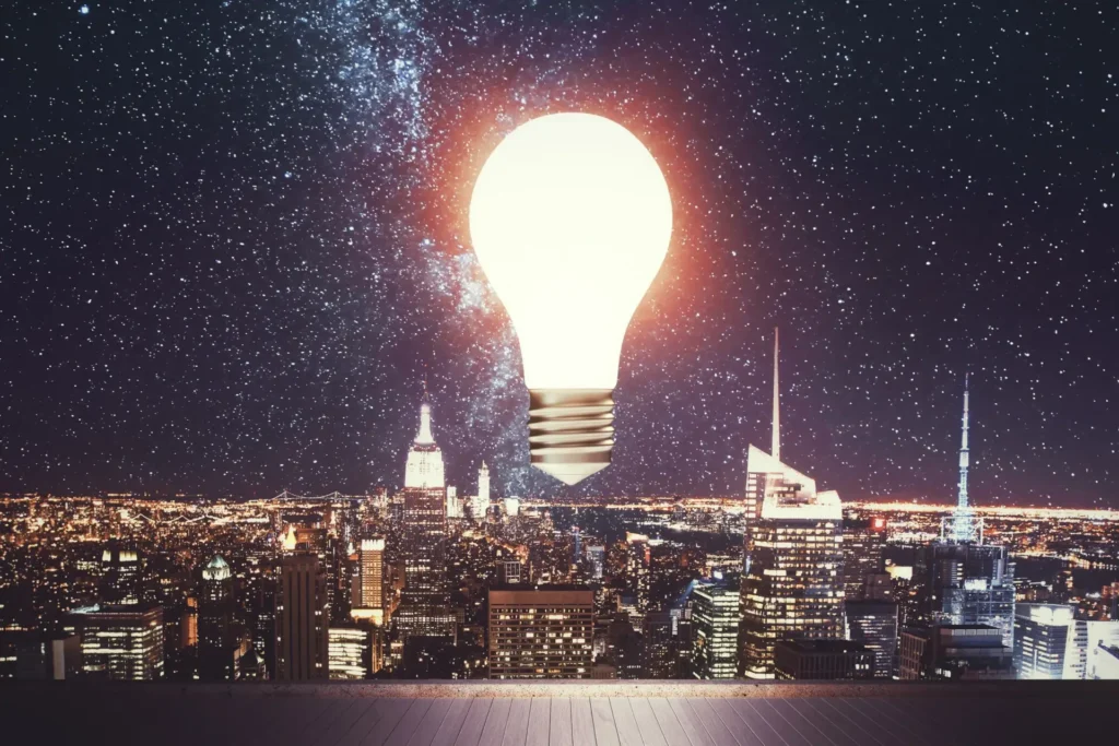 Uma lâmpada brilhante flutua sobre o horizonte noturno de uma cidade brasileira, iluminada por arranha-céus e pela Via Láctea. A imagem simboliza criatividade, inspiração e os desafios da transição energética - valores que a Tecnogera tem orgulho de abraçar.