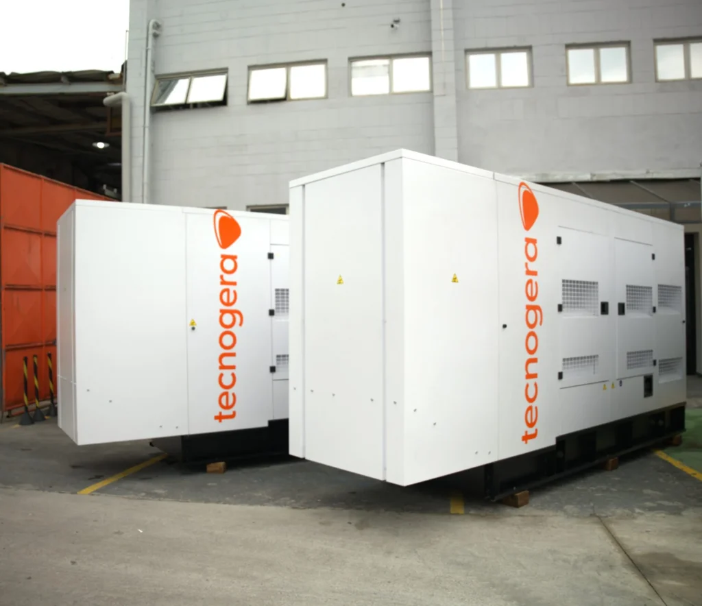 Dois geradores grandes da Tecnogera, estão posicionados sobre paletes ao ar livre, próximos a um prédio cinza. Perfeitos para garantir operações eficientes.