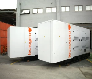 Dois geradores grandes da Tecnogera, estão posicionados sobre paletes ao ar livre, próximos a um prédio cinza. Perfeitos para garantir operações eficientes.