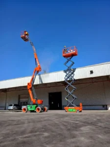 Dois operadores profissionais de plataformas elevatórias da Tecnogera trabalham em frente a um galpão industrial. Ao fundo, é possível ver o céu azul.