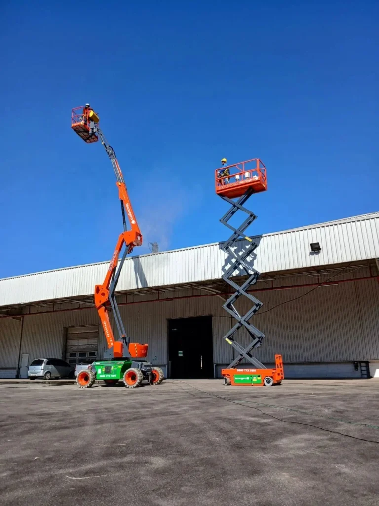 Dois operadores profissionais de plataformas elevatórias da Tecnogera trabalham em frente a um galpão industrial. Ao fundo, é possível ver o céu azul.
