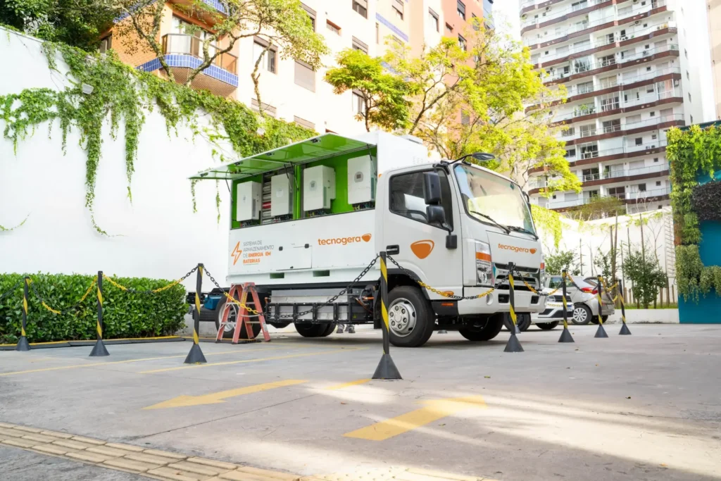 BESS da Tecnogera com detalhes verdes, aberto e carregando baterias em área urbana ensolarada, próximo a prédios e árvores.