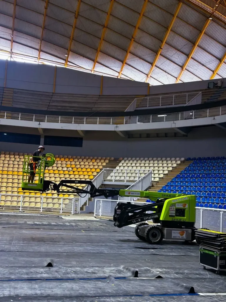Profissional com capacete na plataforma PTA verde Tecnogera, trabalhando no alto de arena coberta, piso protegido por lonas pretas.