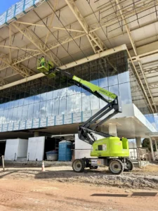 O Equipamento Tecnogera eleva dois profissionais até o teto de obra moderna, com painéis de vidro; materiais ao redor no chão.