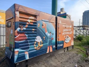 Gerador Tecnogera com ilustrações de trabalhadores e ferramentas, ao ar livre, frase "ENERGÍA QUE MUEVE" e cenário urbano no Oktoberfest Blumenau.