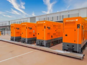 Quatro geradores laranja da Tecnogera, alinhados e prontos para uso em emergências, ao lado de um galpão moderno.