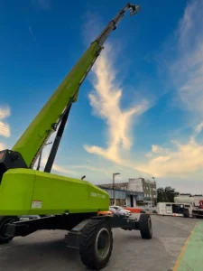 Plataforma elevatória verde da Tecnogera se destaca em altura, sob céu azul, ao lado de veículos e galpões industriais.