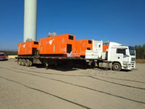 O Caminhão branco Tecnogera transporta quatro geradores laranja sobre o pátio, com base de torre e árvores ao fundo.