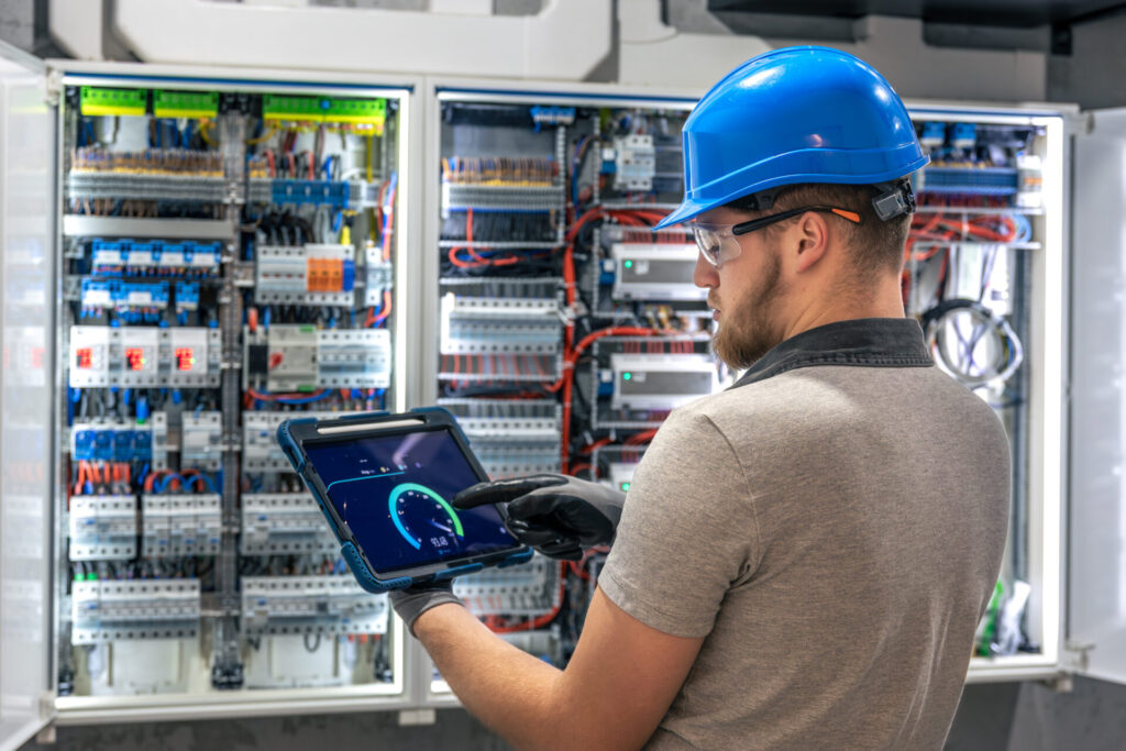 Um técnico da Tecnogera, com capacete azul e tablet, monitora painéis elétricos abertos em ambiente industrial.