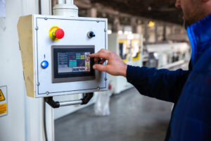 Pessoa de jaqueta azul opera painel touchscreen em equipamento industrial; botões de emergência e reset visíveis.