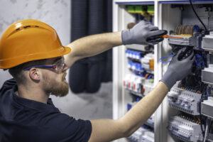 Técnico da Tecnogera com barba, capacete laranja e EPI ajusta fiação em painel elétrico com cabo certificado e disjuntores.