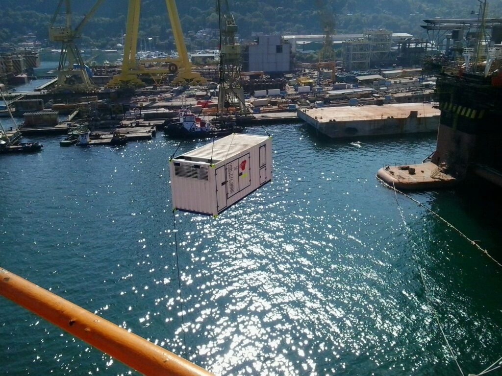 Tecnogera: Um guindaste levanta um contêiner branco sobre a água, com a luz do sol e estruturas industriais ao fundo.